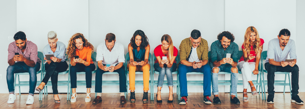 men and women sitting in chairs, lined along a wall, all looking down at their mobile phones