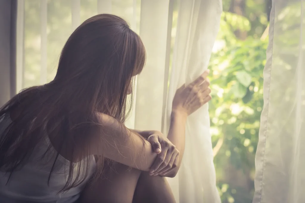 Social anxiety woman nervously peers out the window from behind sheer curtain.